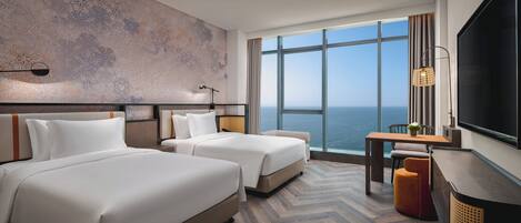 A hotel room with two beds, a large window with a view of the sea, a flat-screen TV, and a wooden herringbone floor.
