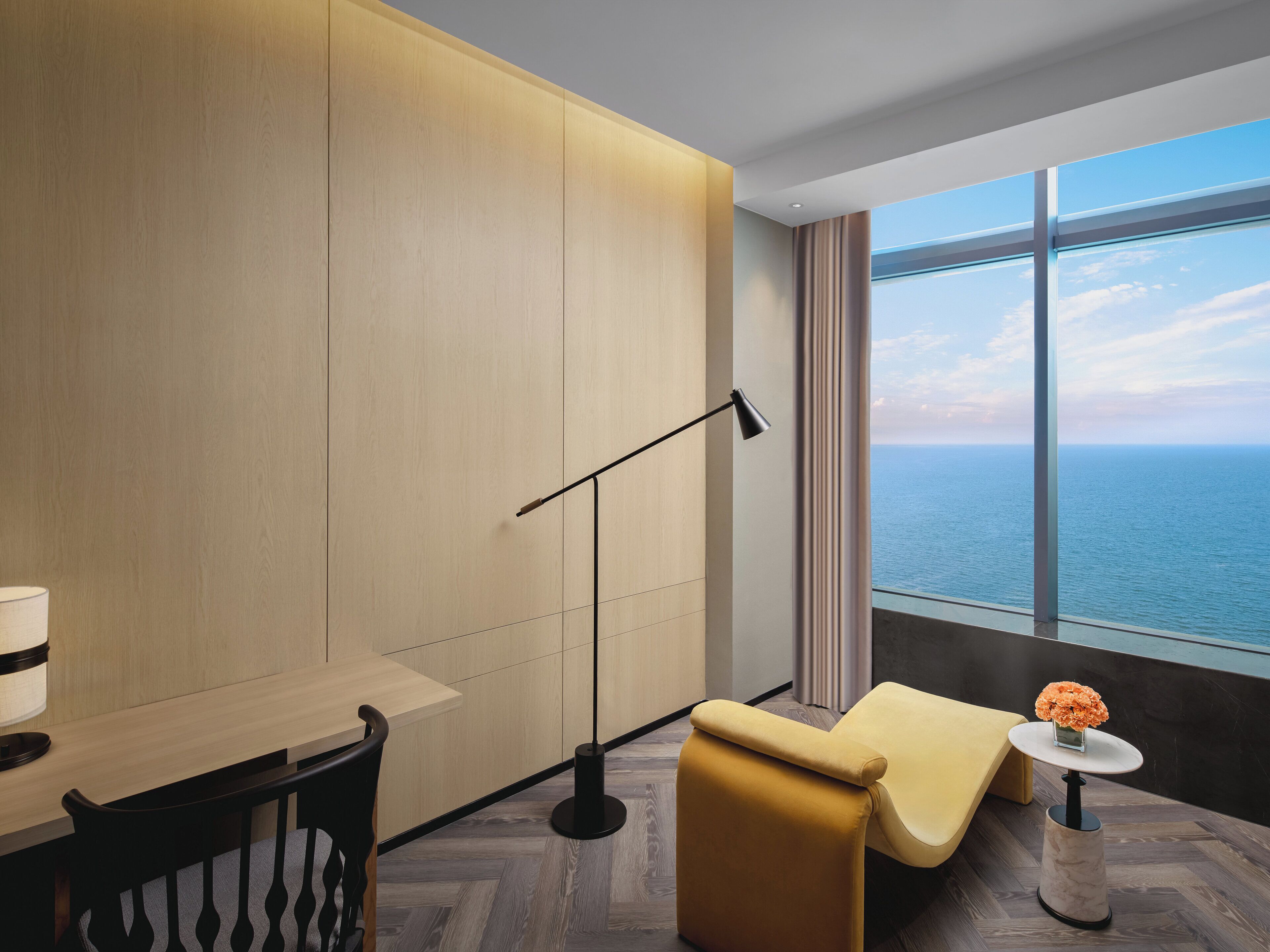 A modern hotel room with a large window offering a view of the sea, a comfortable sofa, a small table with a vase of flowers, and a desk with a lamp.