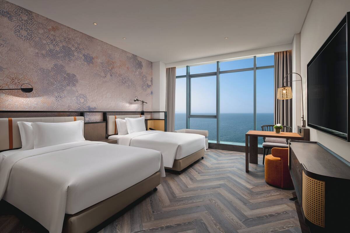 Een hotelkamer met twee bedden, een groot raam met uitzicht op zee, een flatscreen televisie en een houten visgraatvloer.