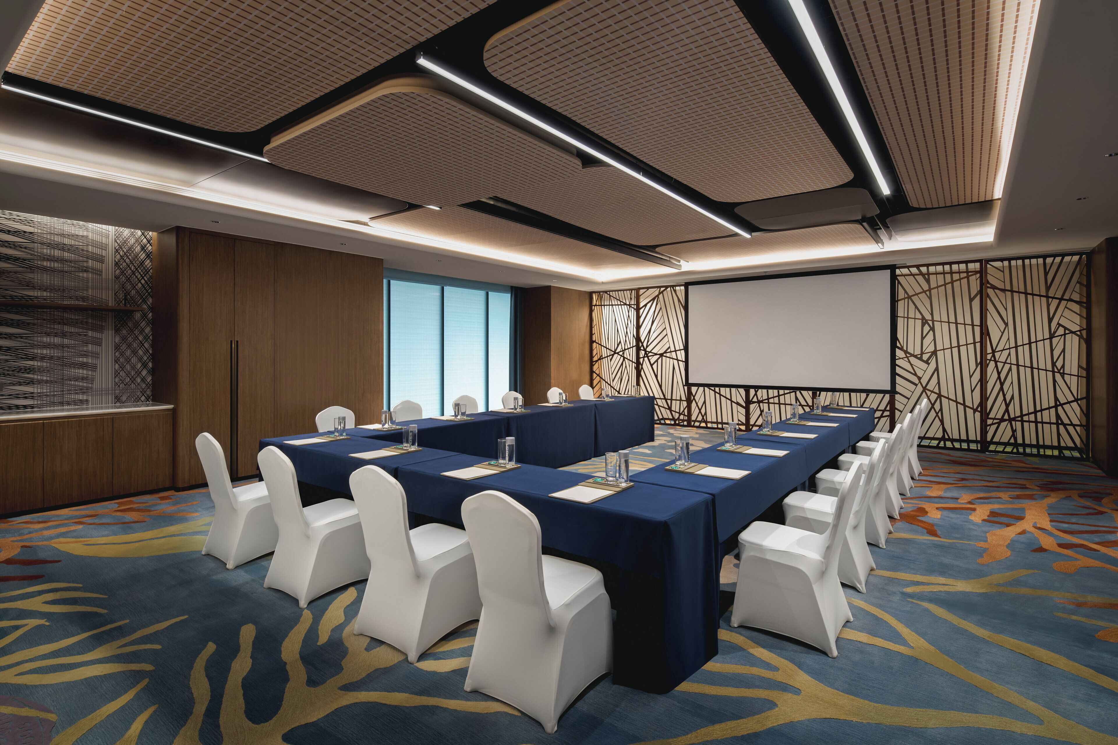 A conference room with a long table, white chairs, and a projector screen.