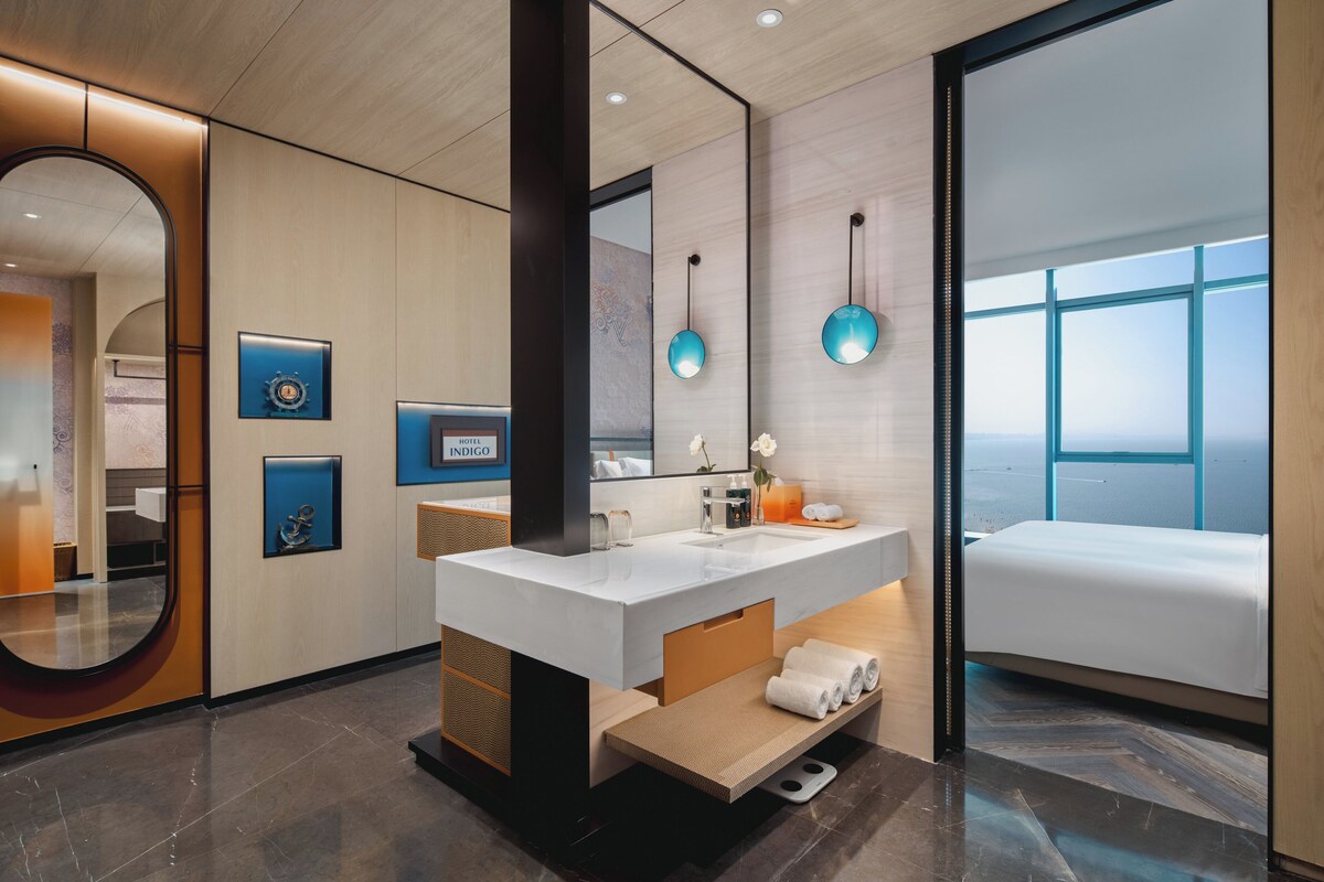 Un baño de hotel moderno con un espejo grande, un lavabo blanco y vistas de la ciudad a través de amplios ventanales.