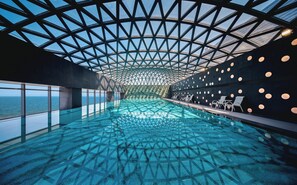 Una piscina interior moderna con techo de cristal geométrico, zona de estar y vistas al mar.