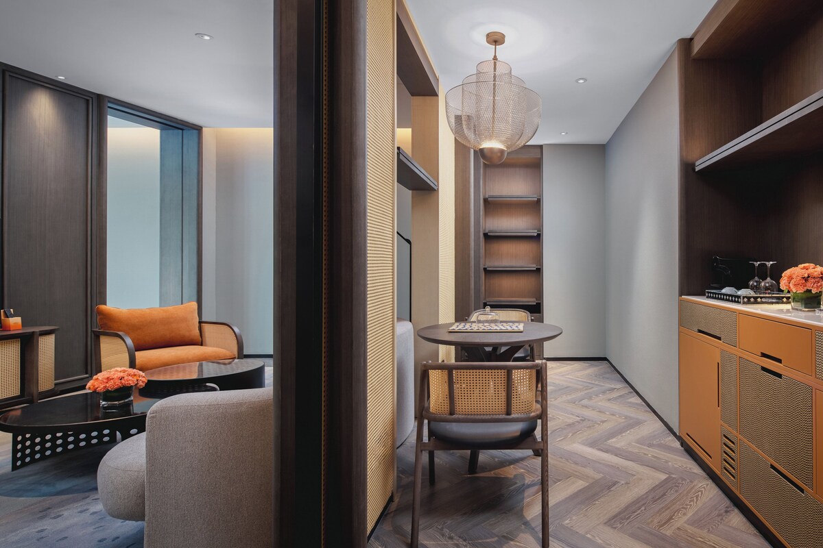 Un interior moderno con una mesa de comedor redonda, una silla de mimbre, un sofá con un cojín naranja y un mueble de madera con estantes.