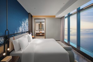 Una habitación de hotel moderna con una cama grande, una mesita de noche, un espejo, un baño con tocador y vistas al océano.