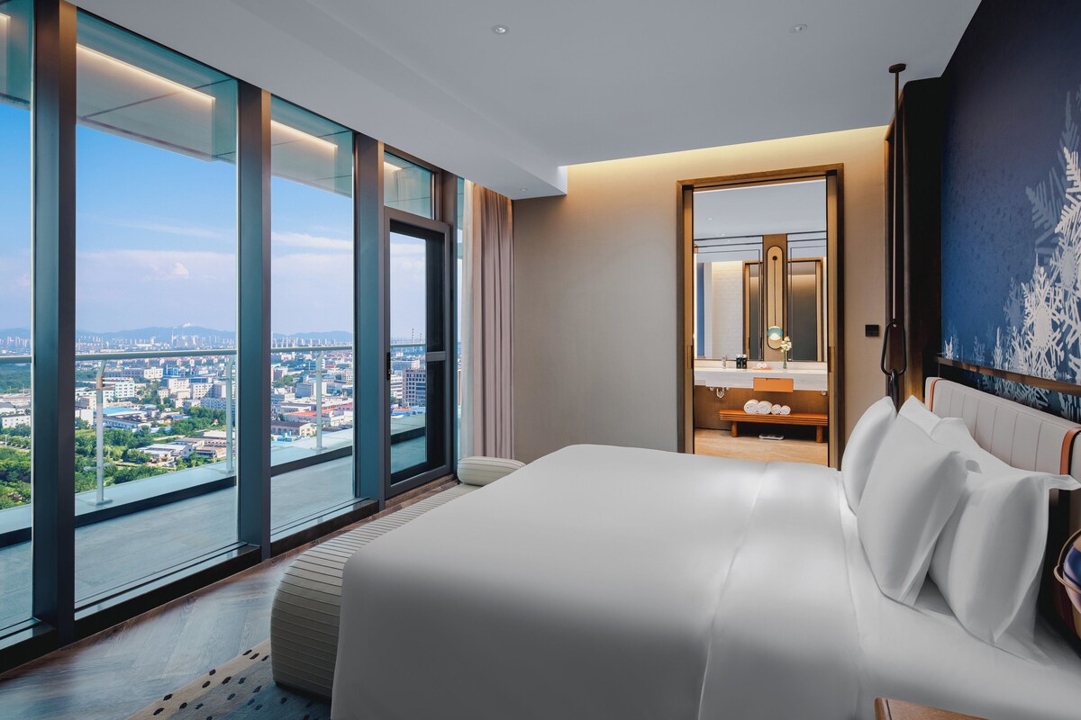 Une chambre d’hôtel moderne dotée d’un grand lit, d’une salle de bain avec un miroir et offrant une vue sur la ville grâce à de grandes fenêtres.