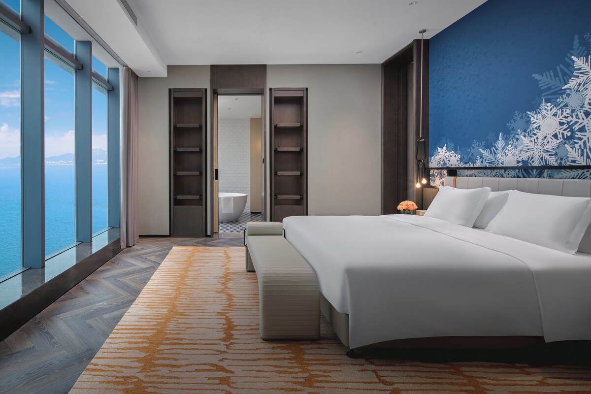 Une chambre d’hôtel moderne dotée d’un grand lit, offrant une vue sur la mer, comprenant une salle de bain avec baignoire et une grande fenêtre.