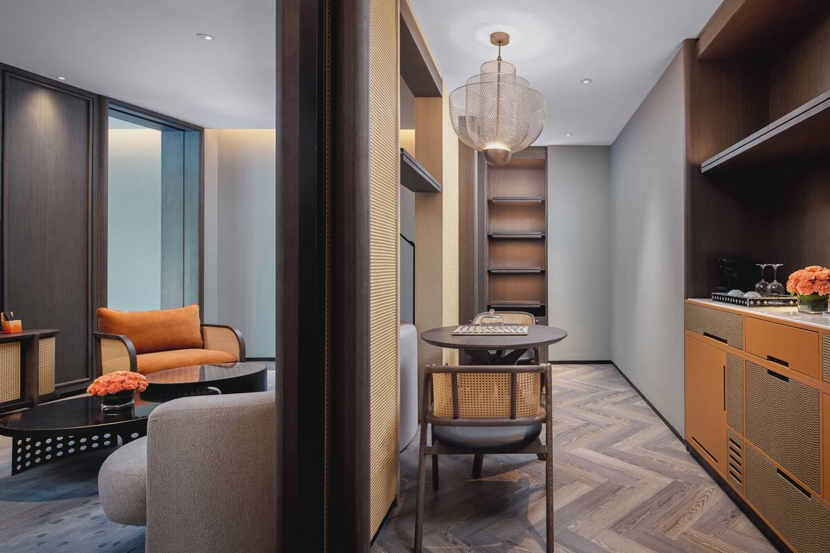Een modern interieur met een ronde eettafel, een rieten stoel, een bank met oranje kussen en een houten kast met planken.