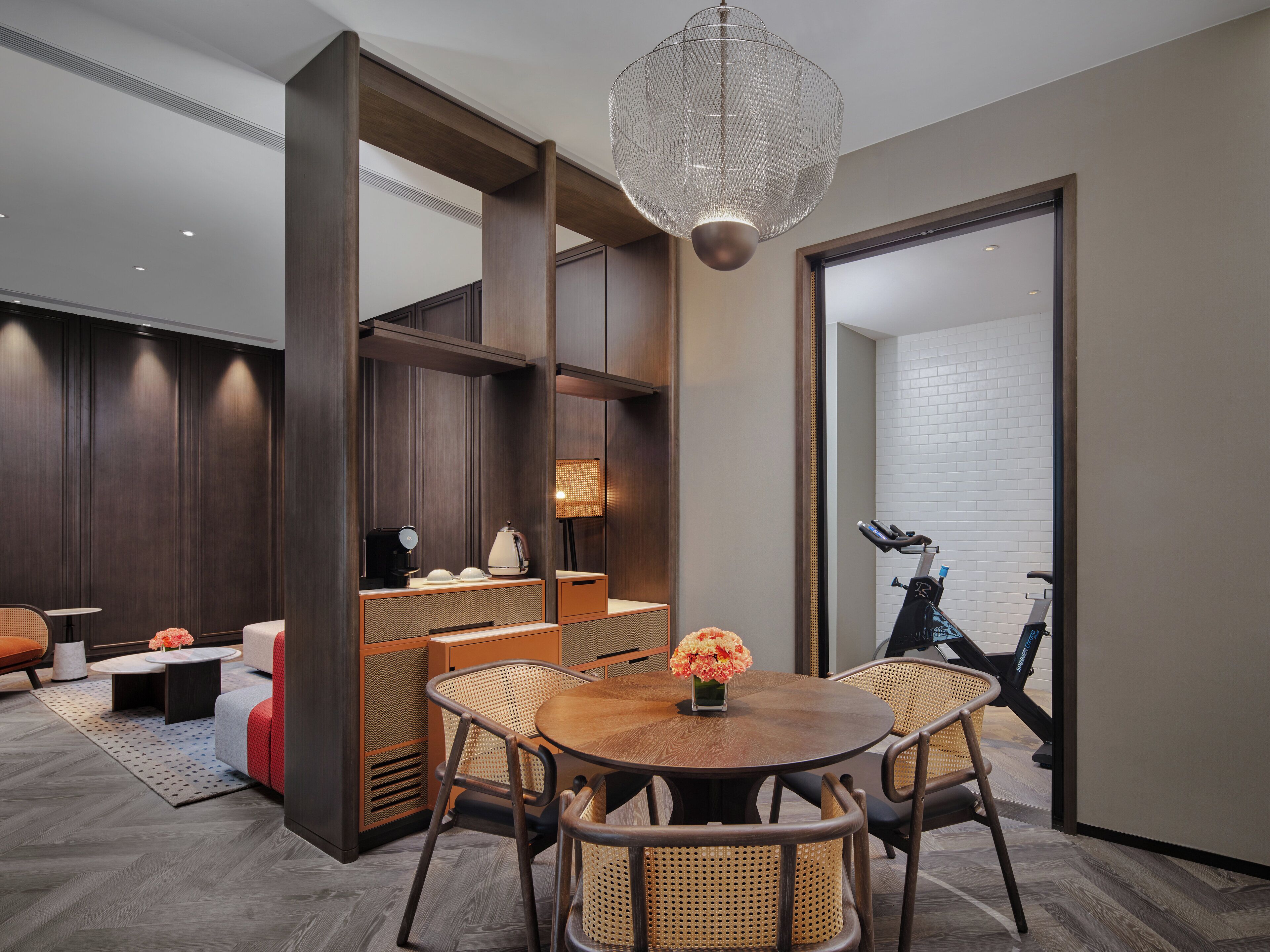 A modern room with a round wooden table, wicker chairs, a coffee station, and a large chandelier.
