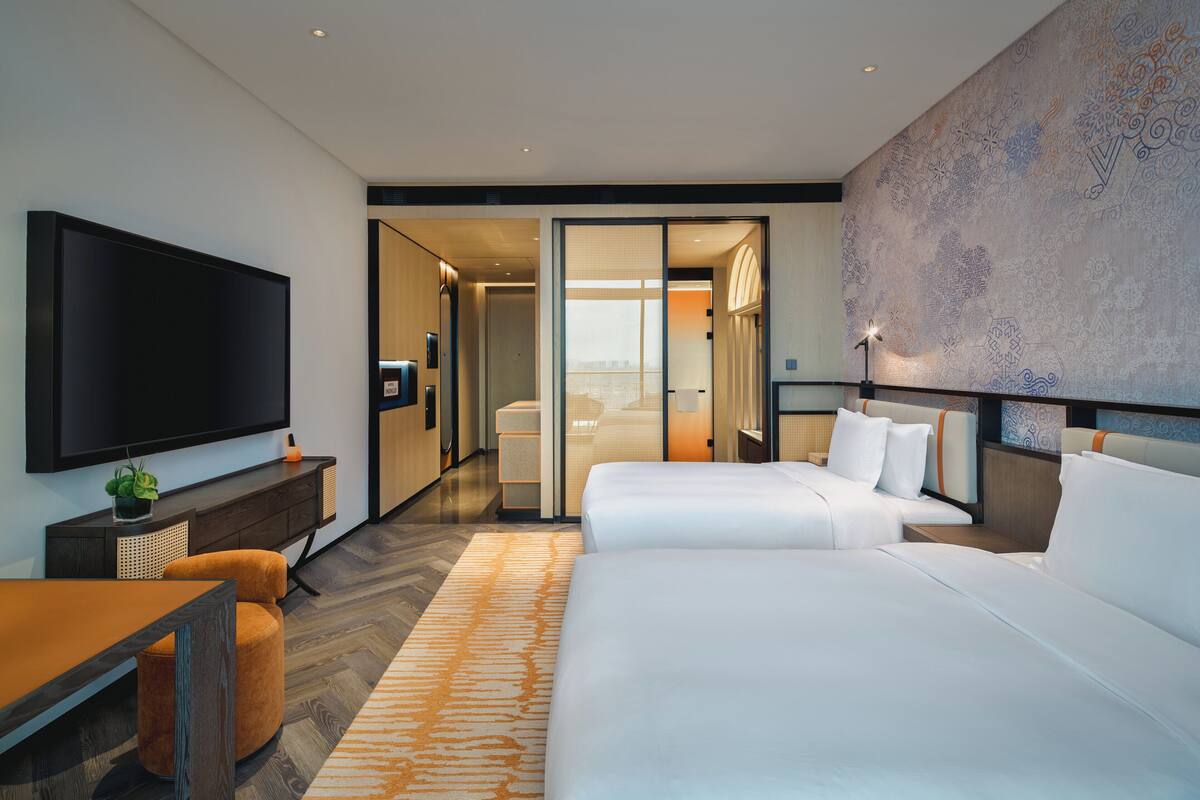 Een moderne hotelkamer met een grote flatscreen-tv, twee bedden, een zithoek en uitzicht op de stad door een schuifdeur.