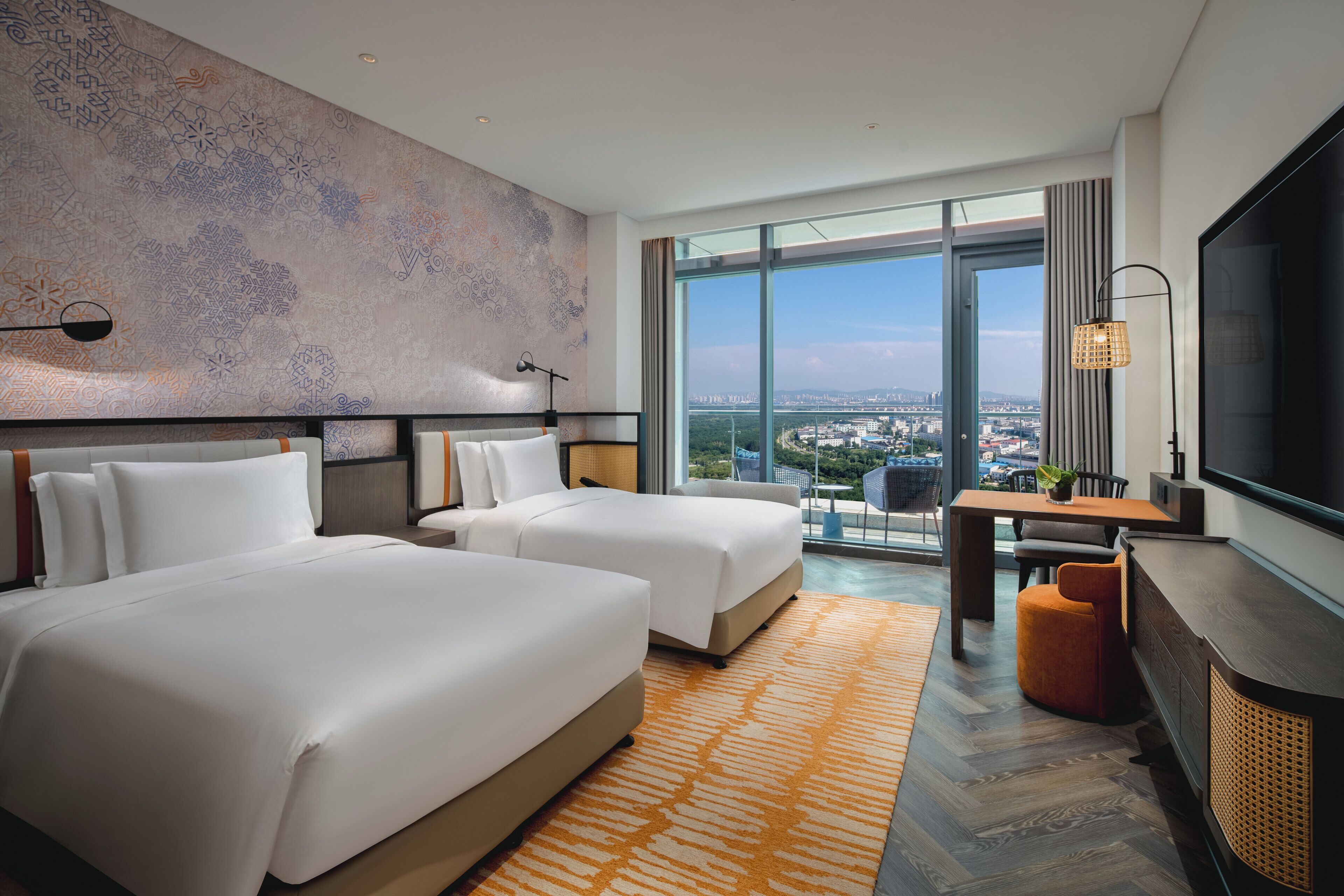 A hotel room with two beds, a desk, a TV, and a large window with a city view.