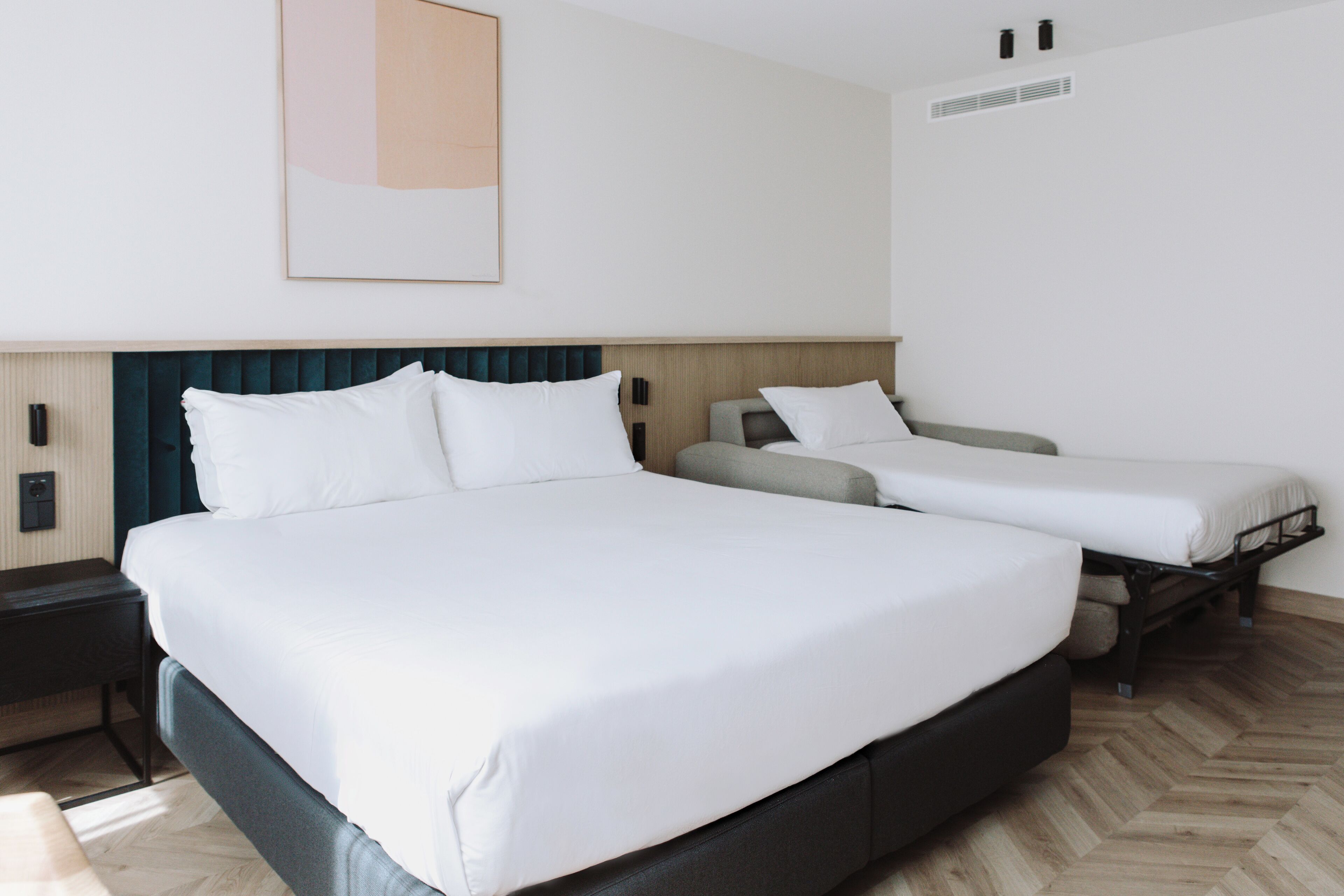 Habitación de hotel con una cama grande, dos camas más pequeñas, una mesita de noche, una obra de arte colgada en la pared y una unidad de ventilación.