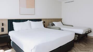 Habitación de hotel con una cama grande, dos camas más pequeñas, una mesita de noche, una obra de arte colgada en la pared y una unidad de ventilación.