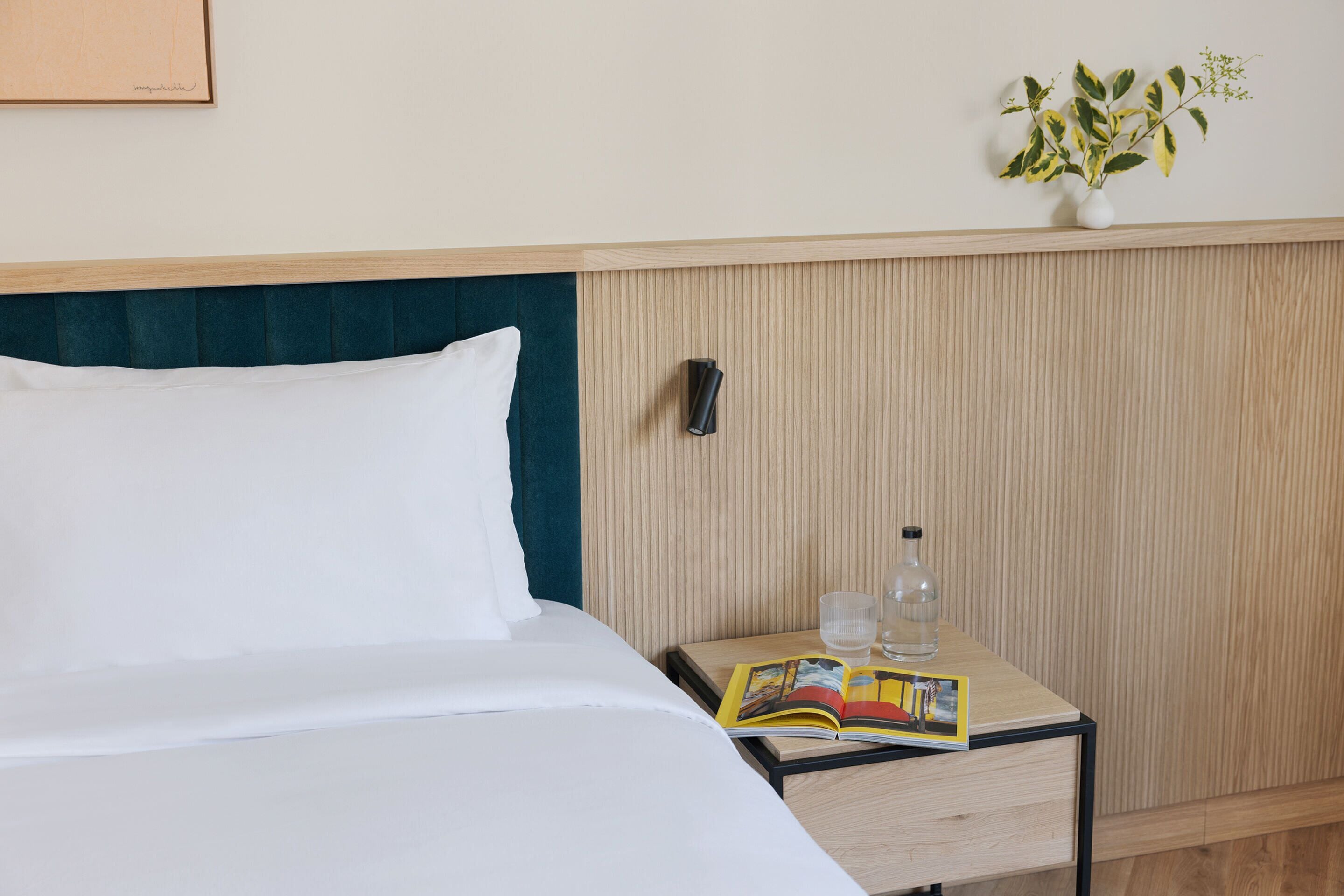 A neatly made bed with white linens, a bedside table with a book, a glass of water, and a vase with a plant.