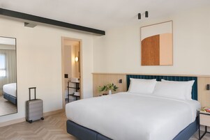 Una habitación de hotel con una cama grande, una mesita de noche, un espejo, una obra de arte colgada en la pared y una maleta.
