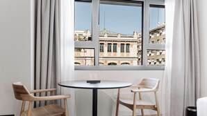 A room with a round table and two wooden chairs, a window with a view of buildings, and a black trash bin. - Aspasios Sofia Apartments (Madrid)