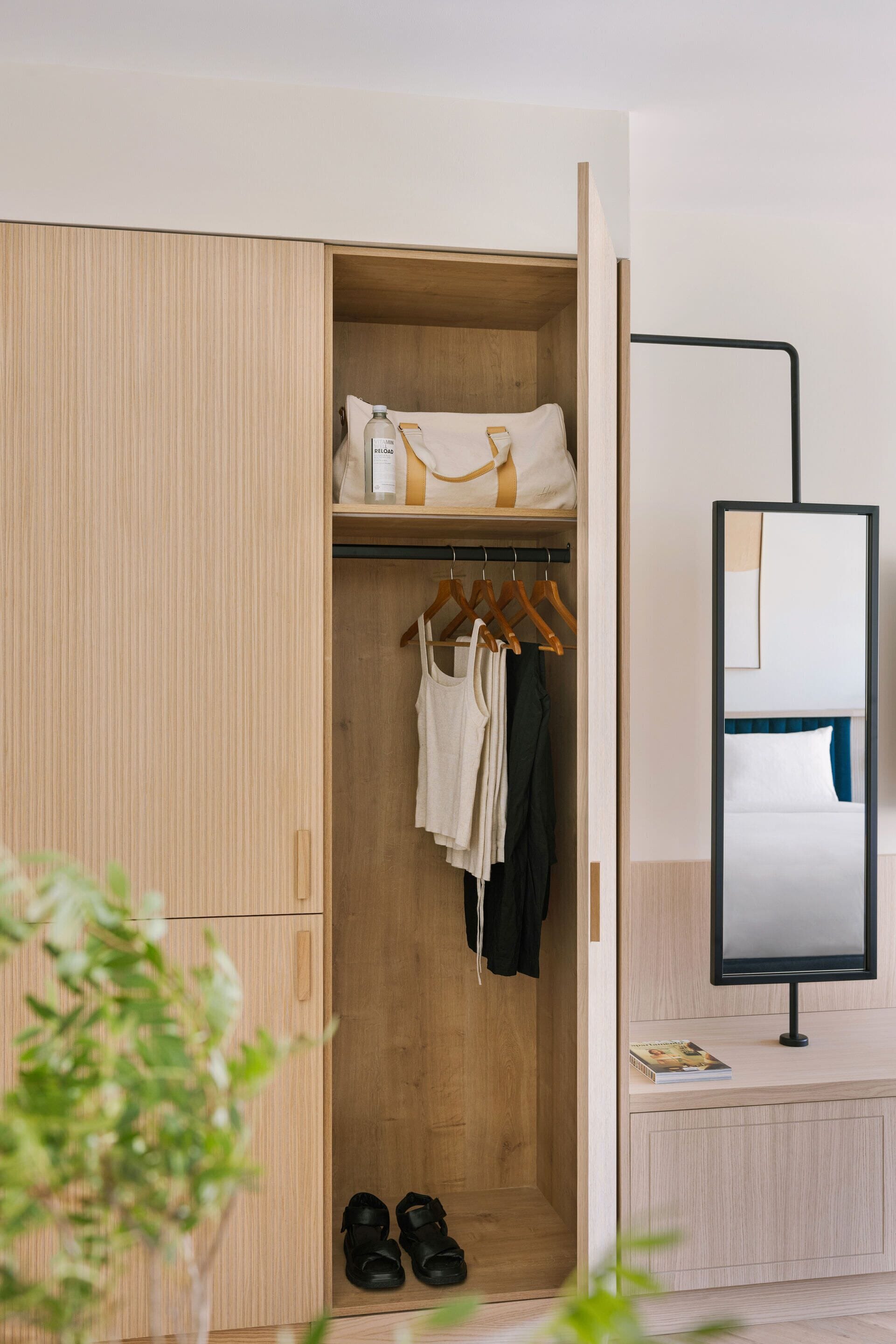 Una habitación con un armario de madera, un perchero, una cama con cabecero y una mesita de noche con una revista.