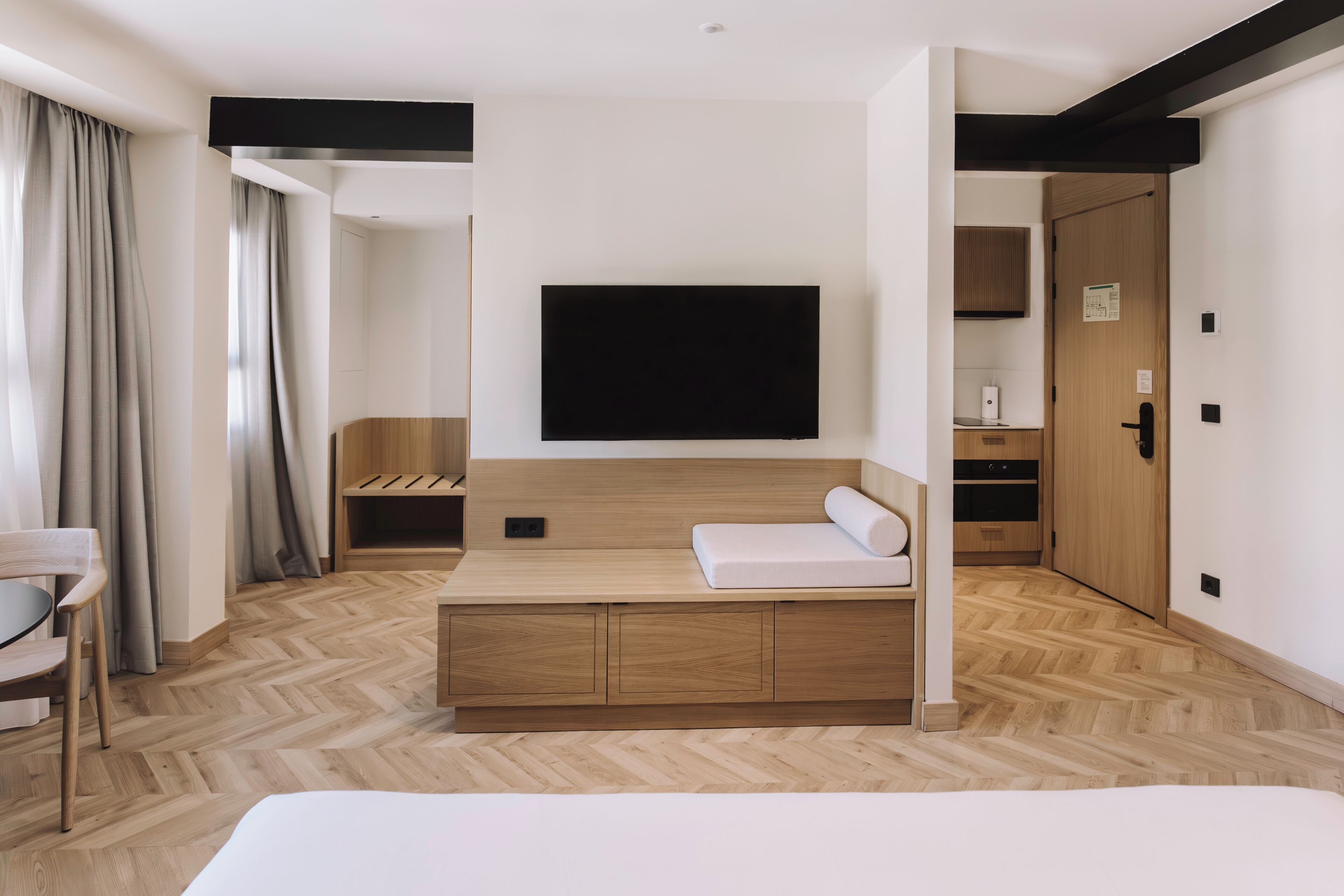 A modern hotel room with a wooden floor, a bed, a TV, a desk, and a chair.