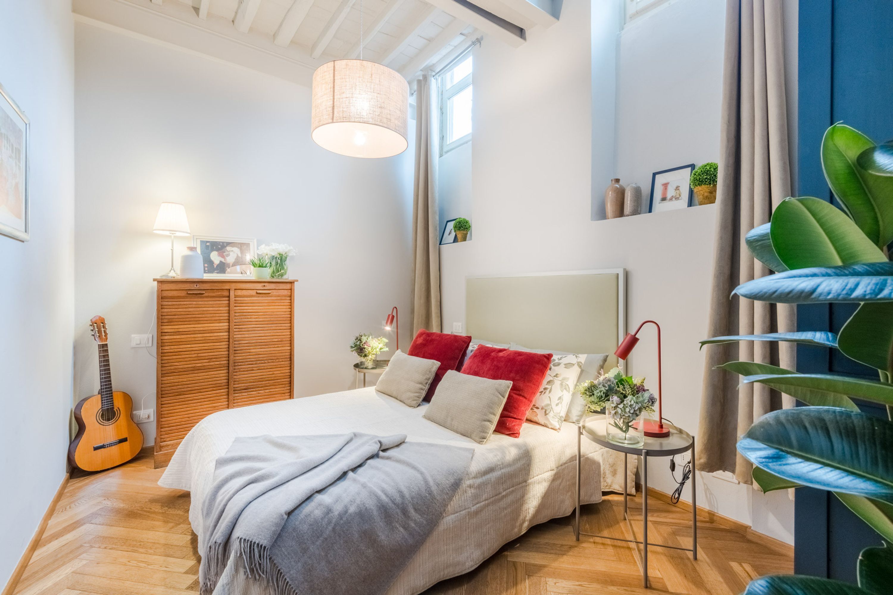 A bedroom with a bed, a guitar, a bedside table with a lamp, and a large plant.