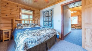 Un dormitorio de cabaña de madera con una cama, un ventilador de techo, una ventana con cortinas y una puerta que da a otra habitación.