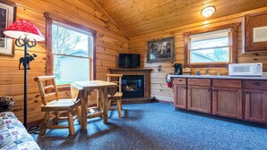 Interior acogedor de una cabaña con piso de madera, chimenea, mesa de comedor con sillas, televisor y zona de cocina con armarios y microondas.