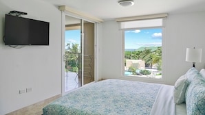 Ein Schlafzimmer mit einem Bett, einem an der Wand montierten Fernseher und einem Fenster mit Blick auf Palmen und einen Pool.