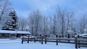 A snow-covered outdoor area with a wooden fence, a building with a snow-covered roof, and trees in the background. - Perfect for Couples, Pet Friendly, Bear Theme, Log Cabin, Fireplace (Mountain)