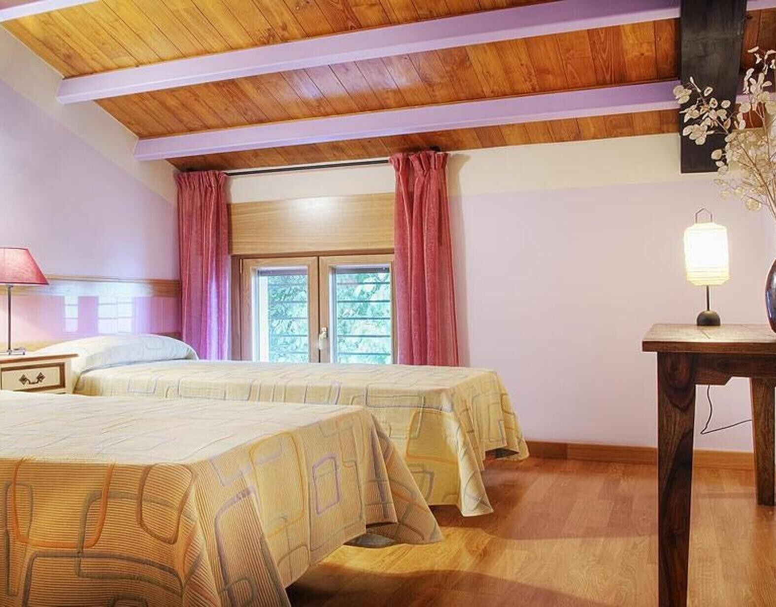 Ein Schlafzimmer mit zwei Betten, Holzbalkendecke, einem Fenster mit Vorhängen, einem Nachttisch mit Lampe und einem kleinen Tisch ebenfalls mit Lampe.