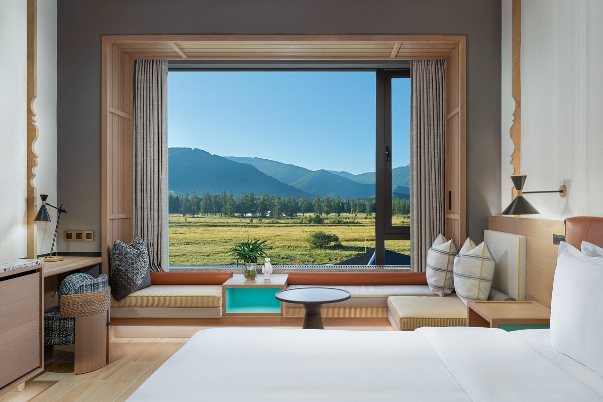 A hotel room with a large window offering a mountain view, a bed, a small table, and a seating area with cushions.