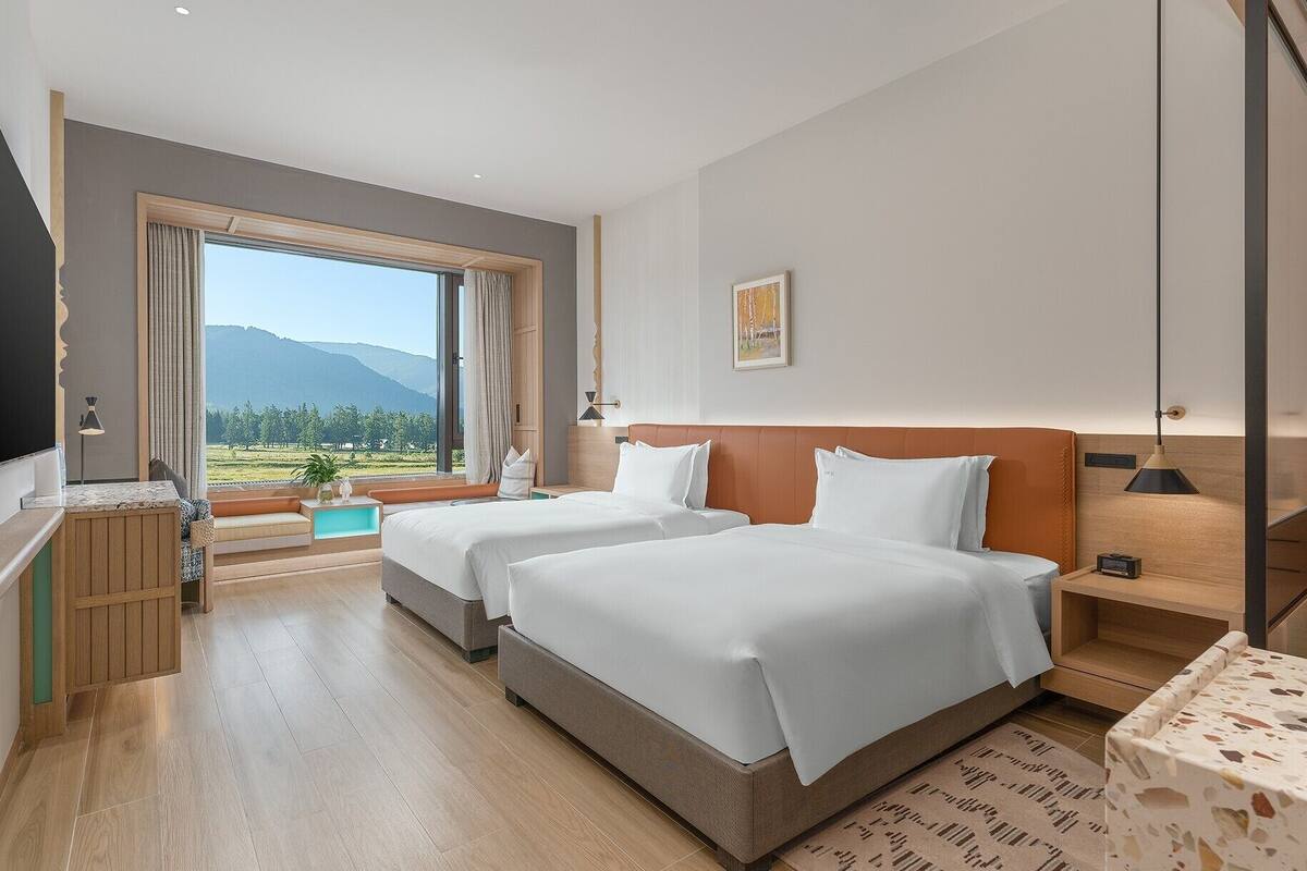 A hotel room with two beds, a large window with a mountain view of mountains, a desk with a lamp, and a small table with a lamp.