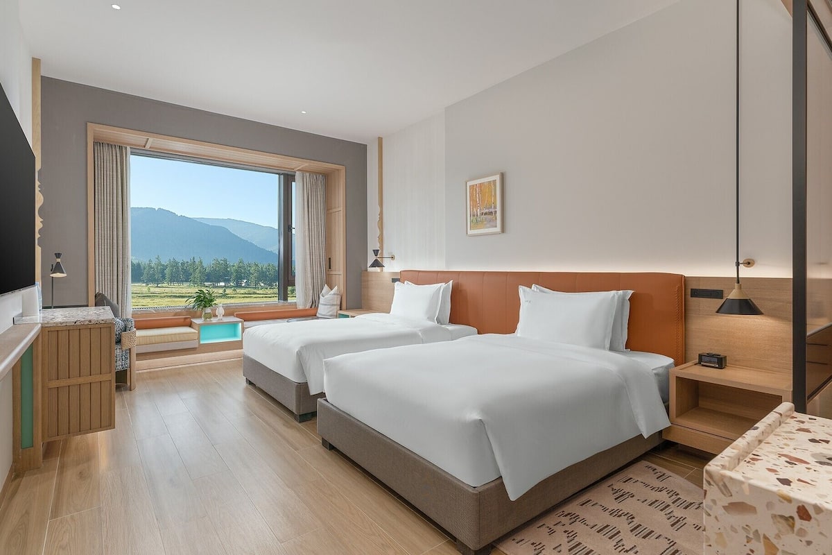 A hotel room with two beds, a large window with a mountain view of mountains, a desk with a lamp, and a small table with a lamp.