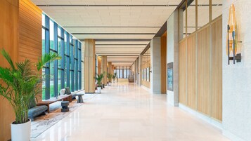 Un lobby de hotel moderno con un largo pasillo, paneles de madera, ventanas amplias y plantas en macetas.