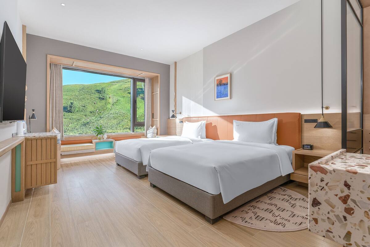 A hotel room with two beds, a TV, a desk, and a window with a view of greenery.
