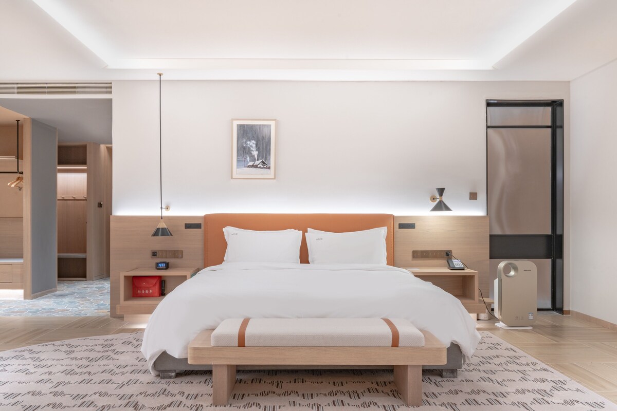 A modern hotel room with a large bed, bedside tables, a bedside lamp, a heater, and a framed picture on the wall.