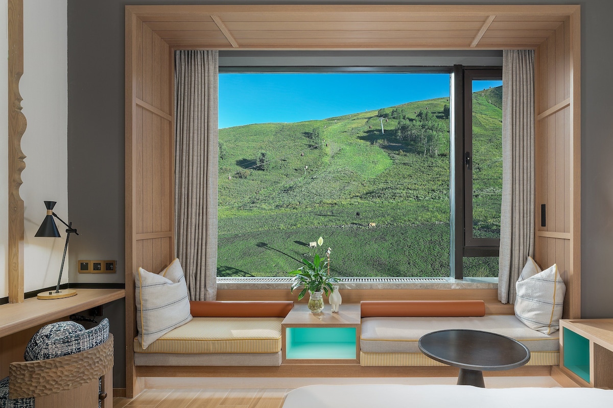 A room with a large window offering a view of a green landscape. There is a bed, a desk, and a chair.