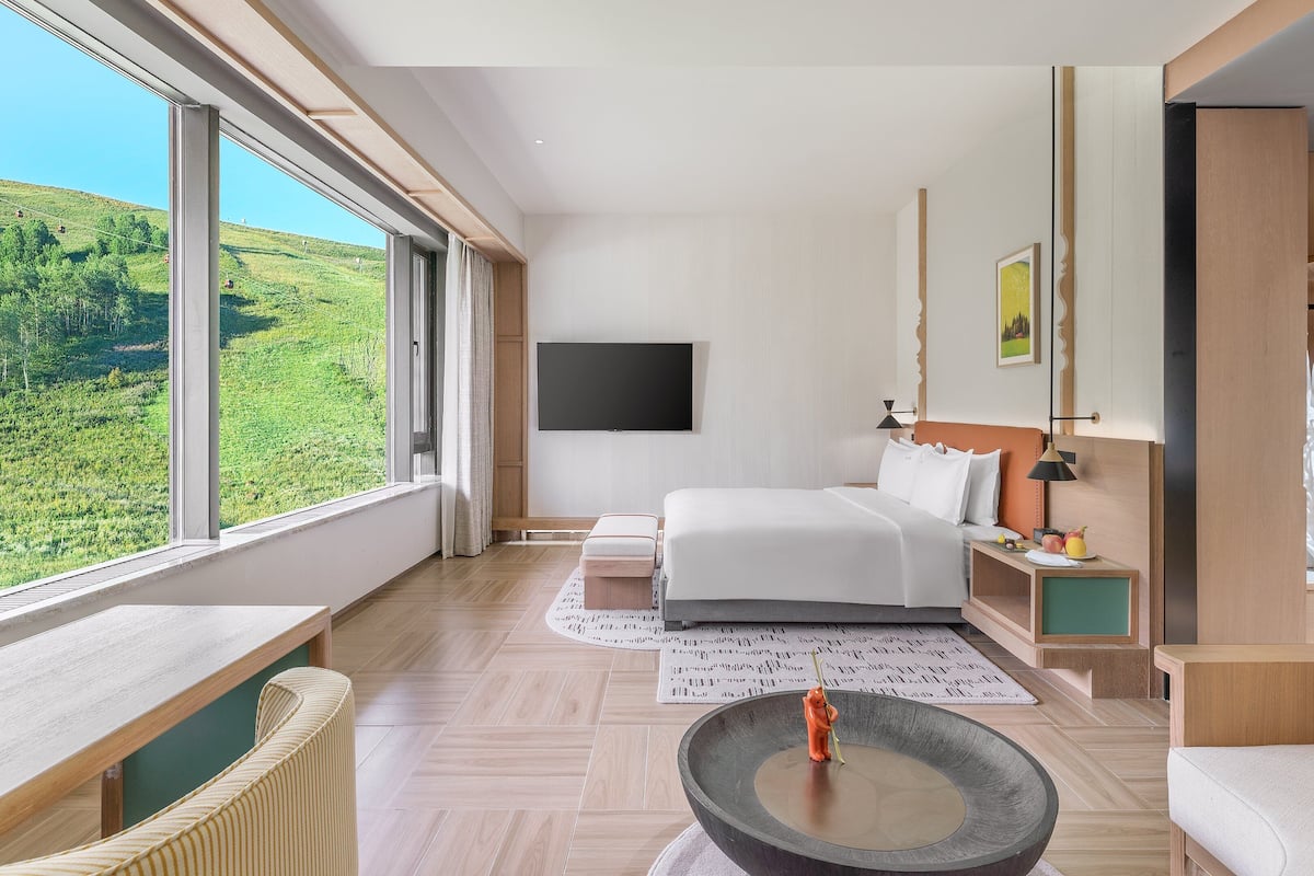 A modern hotel room with a large window offering a scenic view, a bed with white linens, a bedside table with a lamp, and a small seating area with a round table.