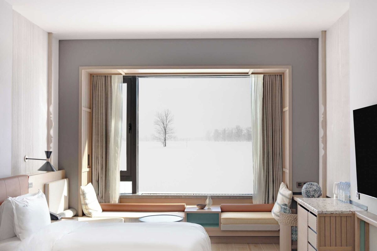 Ein Hotelzimmer mit großem Fenster, einem Bett, einem Schreibtisch und Blick auf eine verschneite Landschaft.
