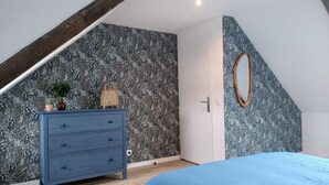 Un dormitorio con techo inclinado, un aparador azul, una cama con colcha azul, un espejo con marco de madera y papel tapiz estampado.