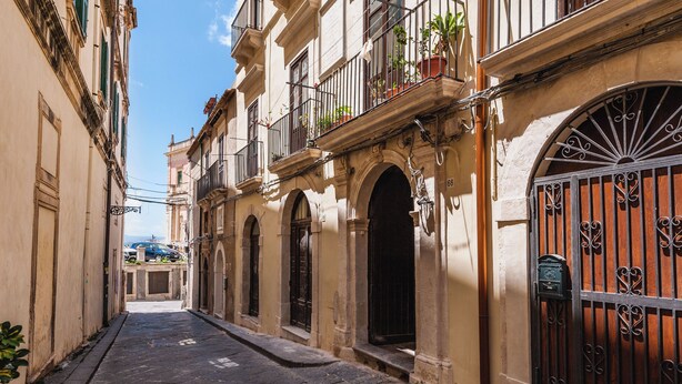 A narrow street with buildings on both sides, featuring balconies with potted plants, arched doorways, and a large wrought iron gate. - Casa Amalfitana by Wonderful Italy (Syracuse)