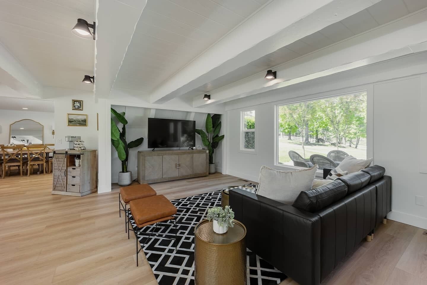 A modern living room with a black leather sofa, wooden dining table, and large windows.