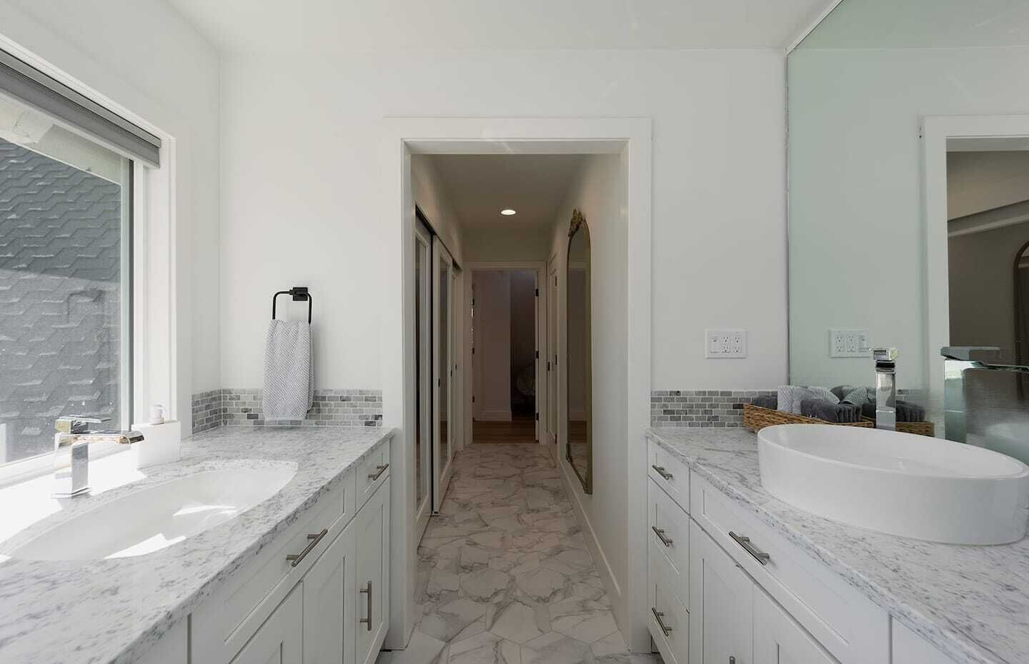 A modern bathroom with two sinks, a large mirror, and a hallway leading to another room.