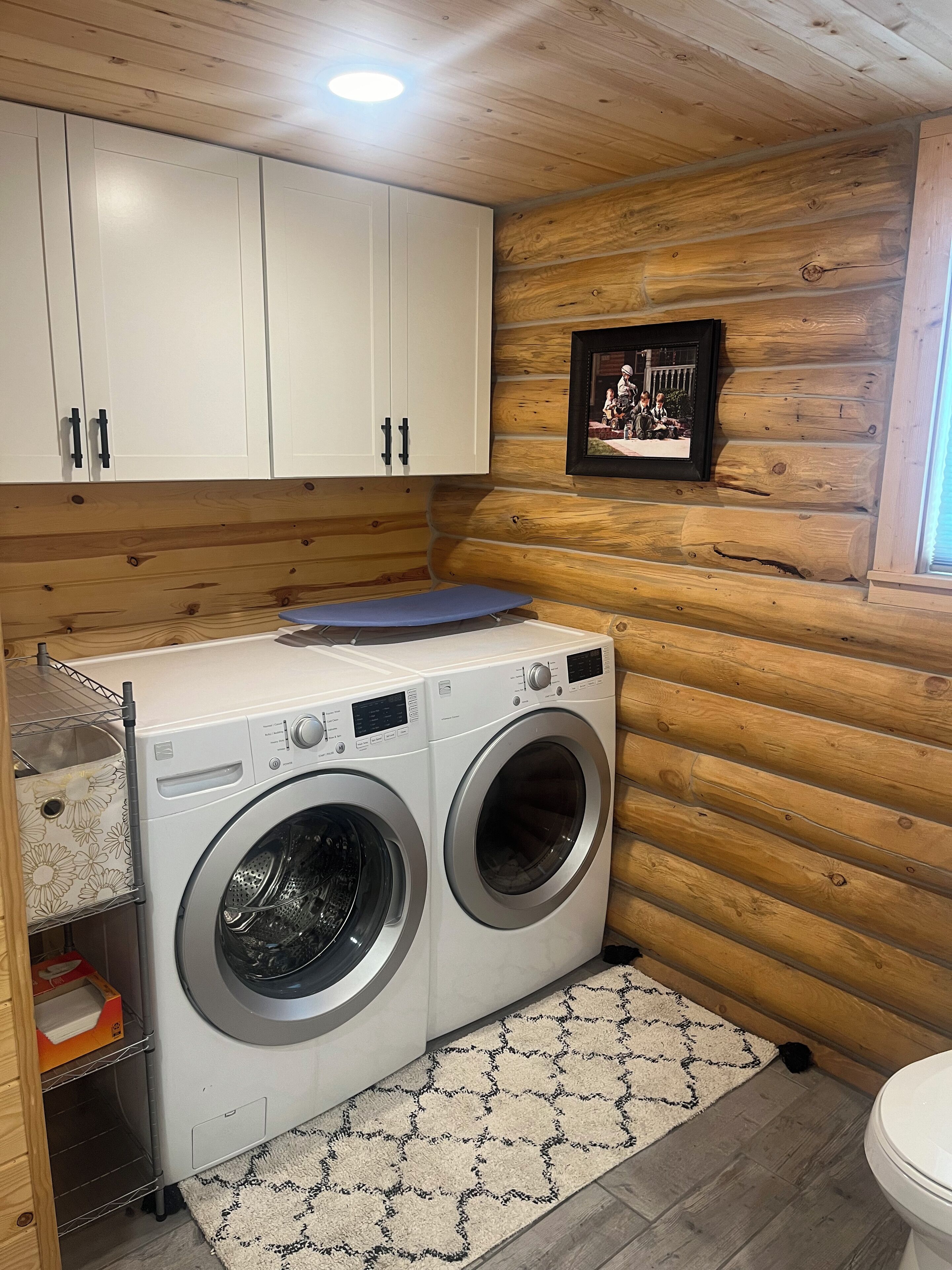 Ein Waschraum mit zwei Waschmaschinen, die von vorne geladen werden, Holzausbau, einem gerahmten Foto an der Wand und einem gemusterten Teppich.