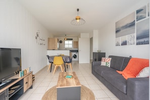 Ein modernes Wohnzimmer mit Flachbildfernseher, Couchtisch aus Holz, grauem Sofa und einem Essbereich mit gelben Stühlen.