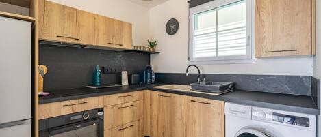 Una cocina moderna con armarios de madera, horno integrado, lavadora y ventana con persianas.