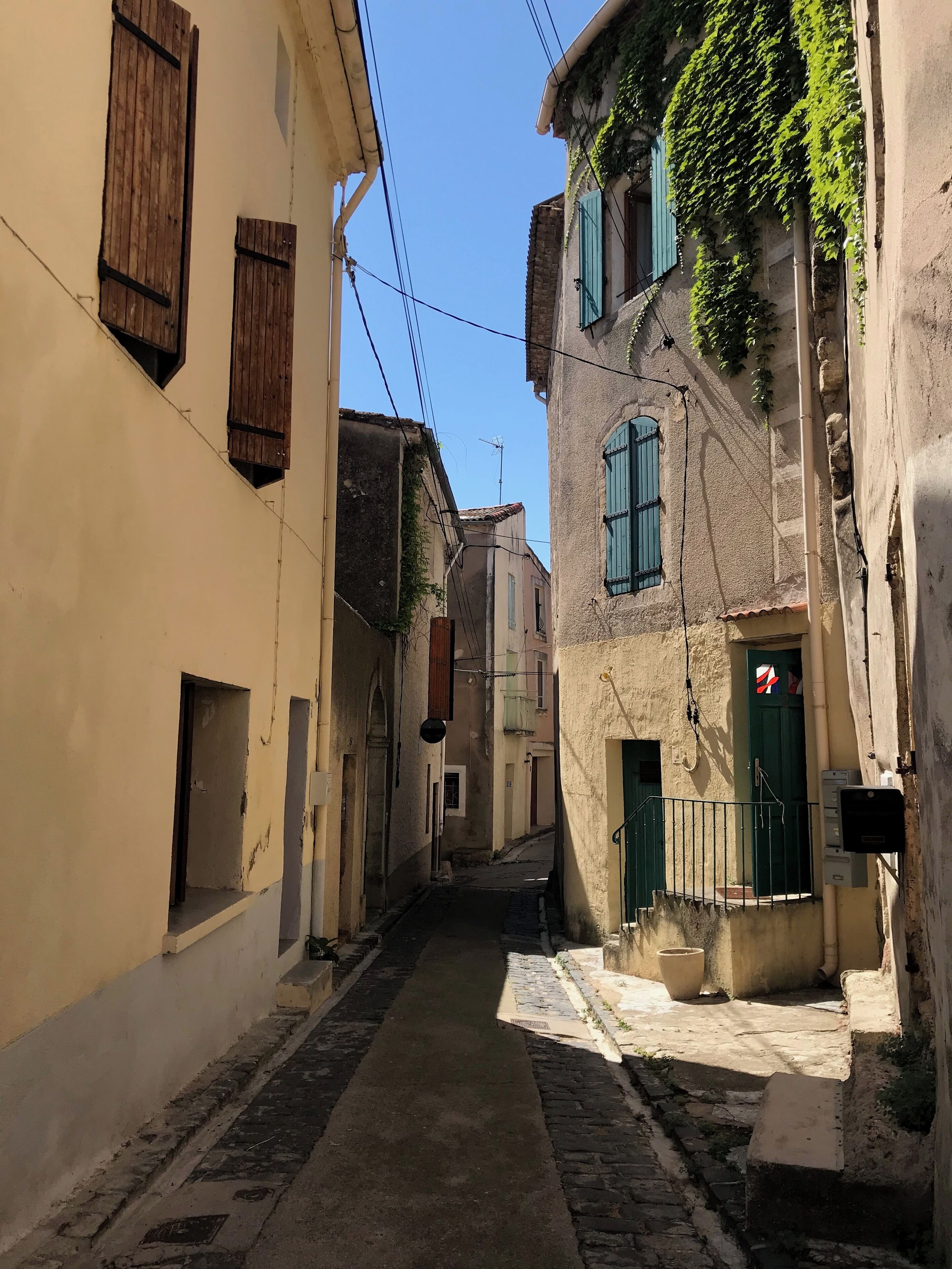 Una stretta strada acciottolata fiancheggiata da edifici con persiane di legno e porte verdi, che evoca un'ambientazione europea storica.
