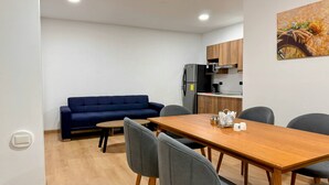 Uma sala de estar moderna com um sofá, uma mesa de jantar com cadeiras, uma área de cozinha com geladeira e armários, e um quadro na parede.