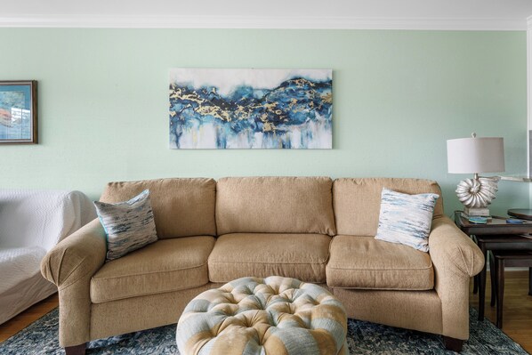 A beige sofa with two matching cushions, a round ottoman, a framed abstract painting, and a lamp on a side table.