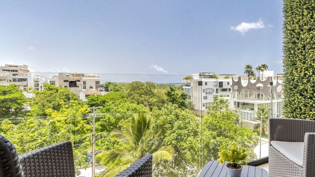 Un balcó amb moble de vimet, una planta en test i vista a edificis moderns i vegetació.