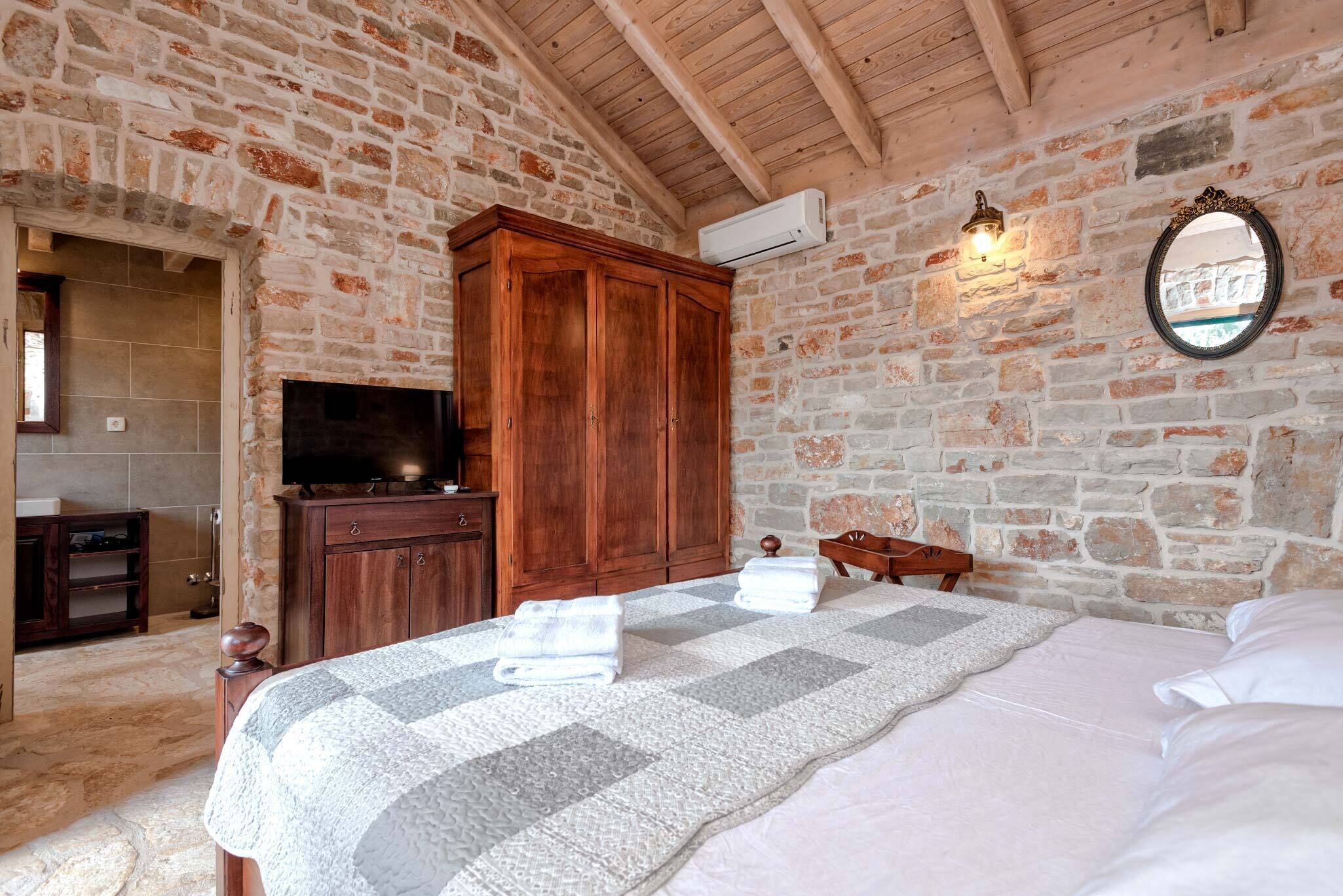 A bedroom with a stone wall, wooden wardrobe, bed with pillows, and a television on a wooden cabinet.