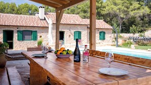 Une table en bois, dressée avec un saladier de fruits, une bouteille et des verres, donnant sur une piscine et des bâtiments en pierre aux volets verts.