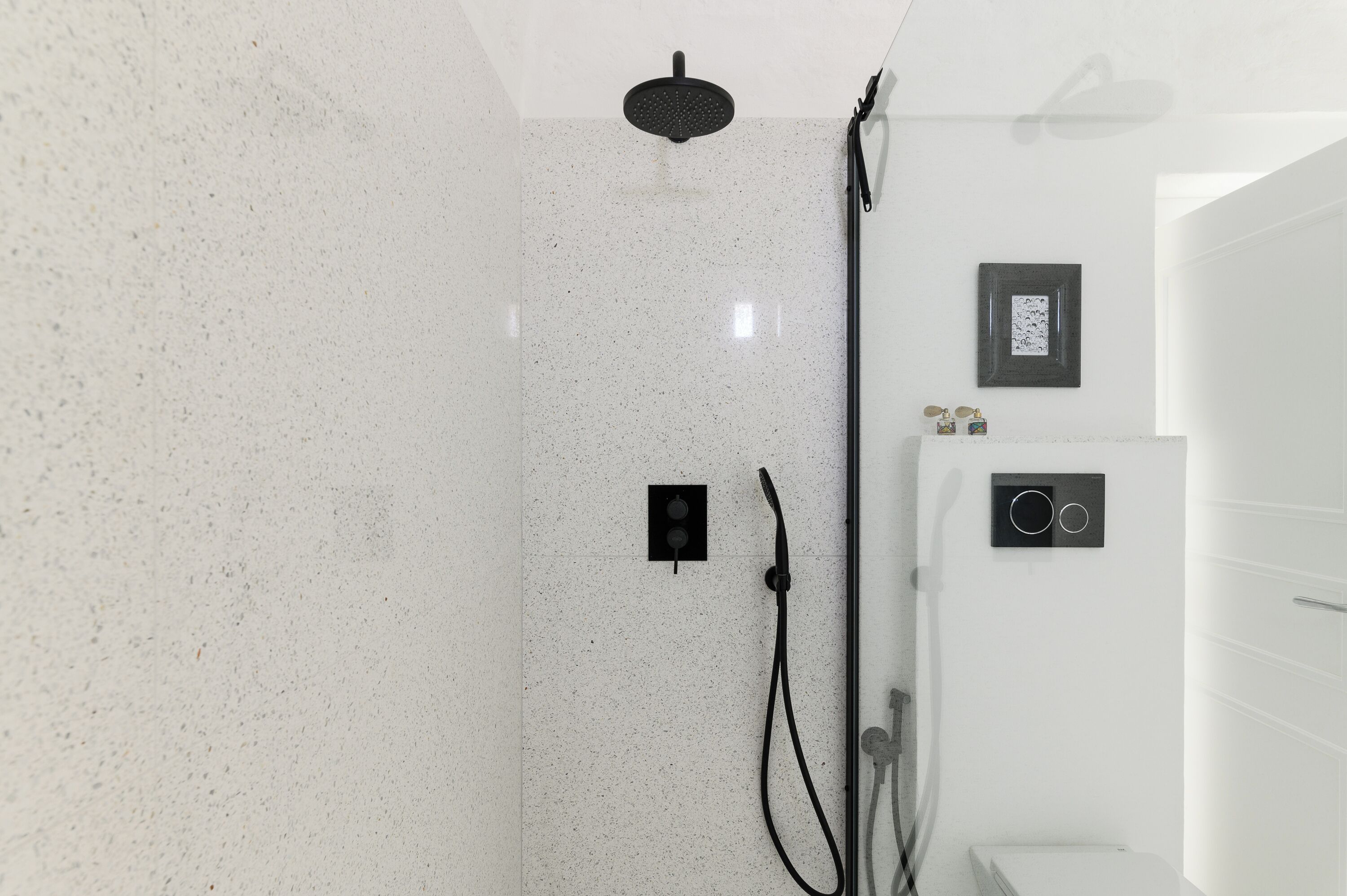 A modern bathroom with a white speckled tile wall, a black showerhead, and a wall-mounted control panel.